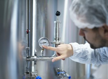 Technologist in white protective suit checking pressure on pressure gauge on industrial machine in factory.